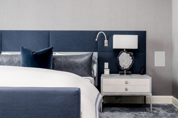 The image shows a modern bedroom with a blue and white color scheme. A bed with blue pillows and a white comforter is next to a nightstand with a lamp.