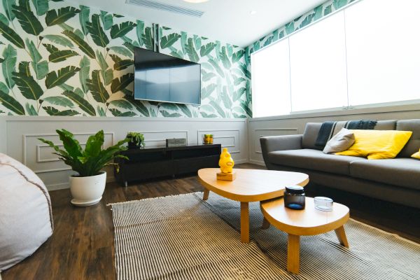 A modern living room with a grey couch, yellow cushions, wooden tables, plant decorations, and patterned wallpaper featuring green leaves.