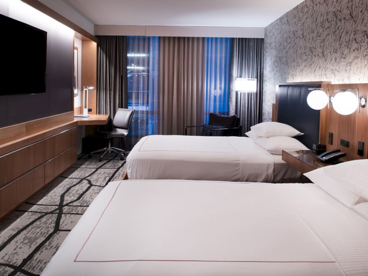 A modern hotel room with two double beds, a wall-mounted TV, a work desk, an armchair, and a well-lit ambiance.