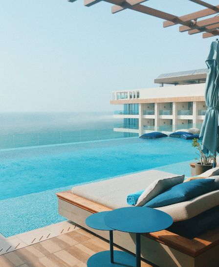A luxurious poolside area with sun loungers, an umbrella, and a view of the sea, surrounded by a modern building.