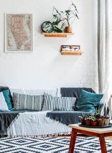 A cozy living room with a sofa, potted plants, a coffee table, and a wall map. Light streams through large windows on the right.