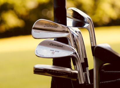 The image shows golf clubs in a golf bag with a blurred green background, showcasing brands like Titleist and Cleveland.