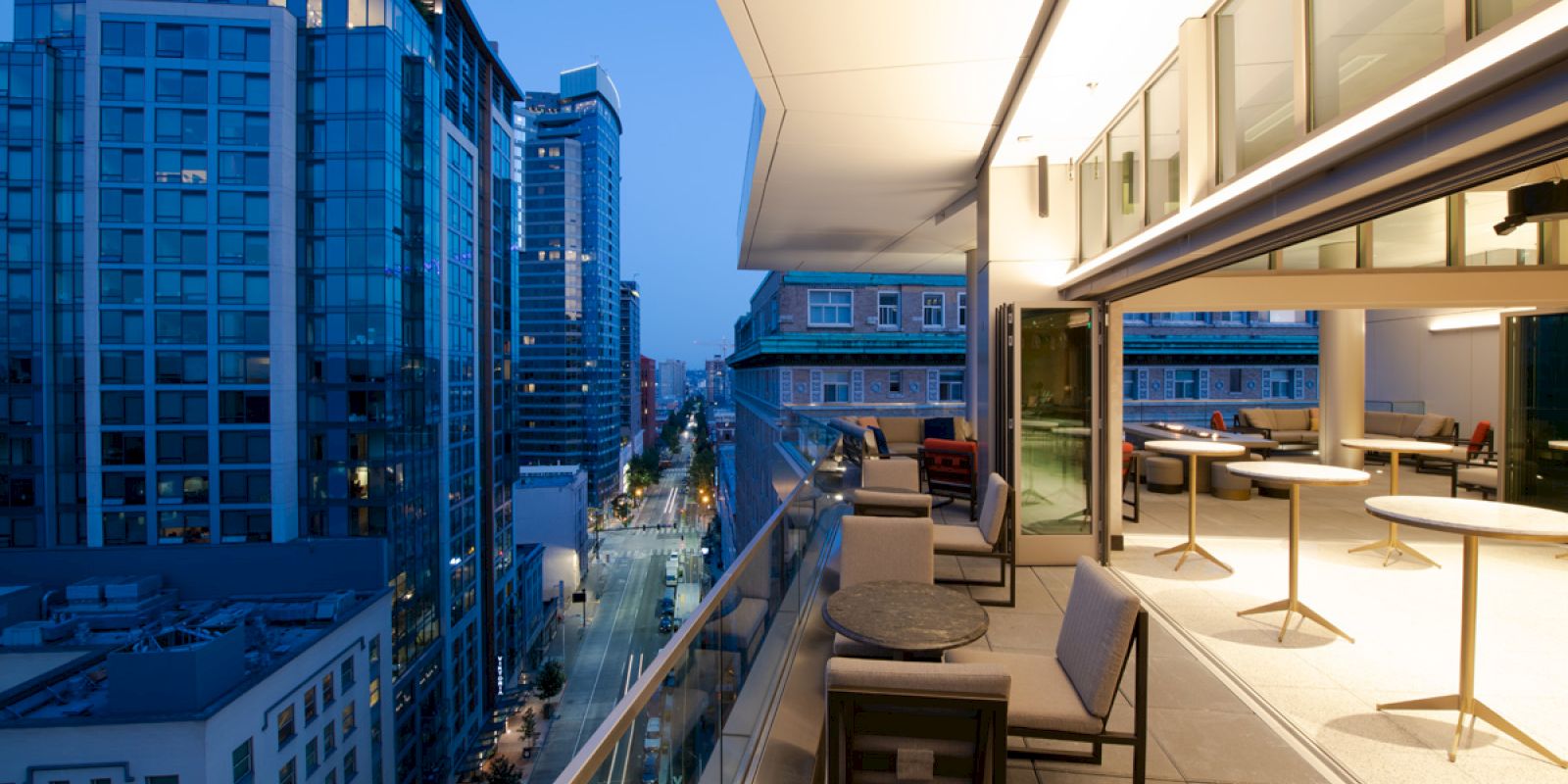 A modern rooftop terrace at sunset, overlooking a cityscape with tall buildings; features seating areas and outdoor tables along a glass railing.