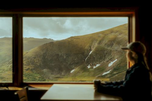 A person sits at a table, looking out a window at a scenic mountain landscape under a cloudy sky, with patches of snow visible.