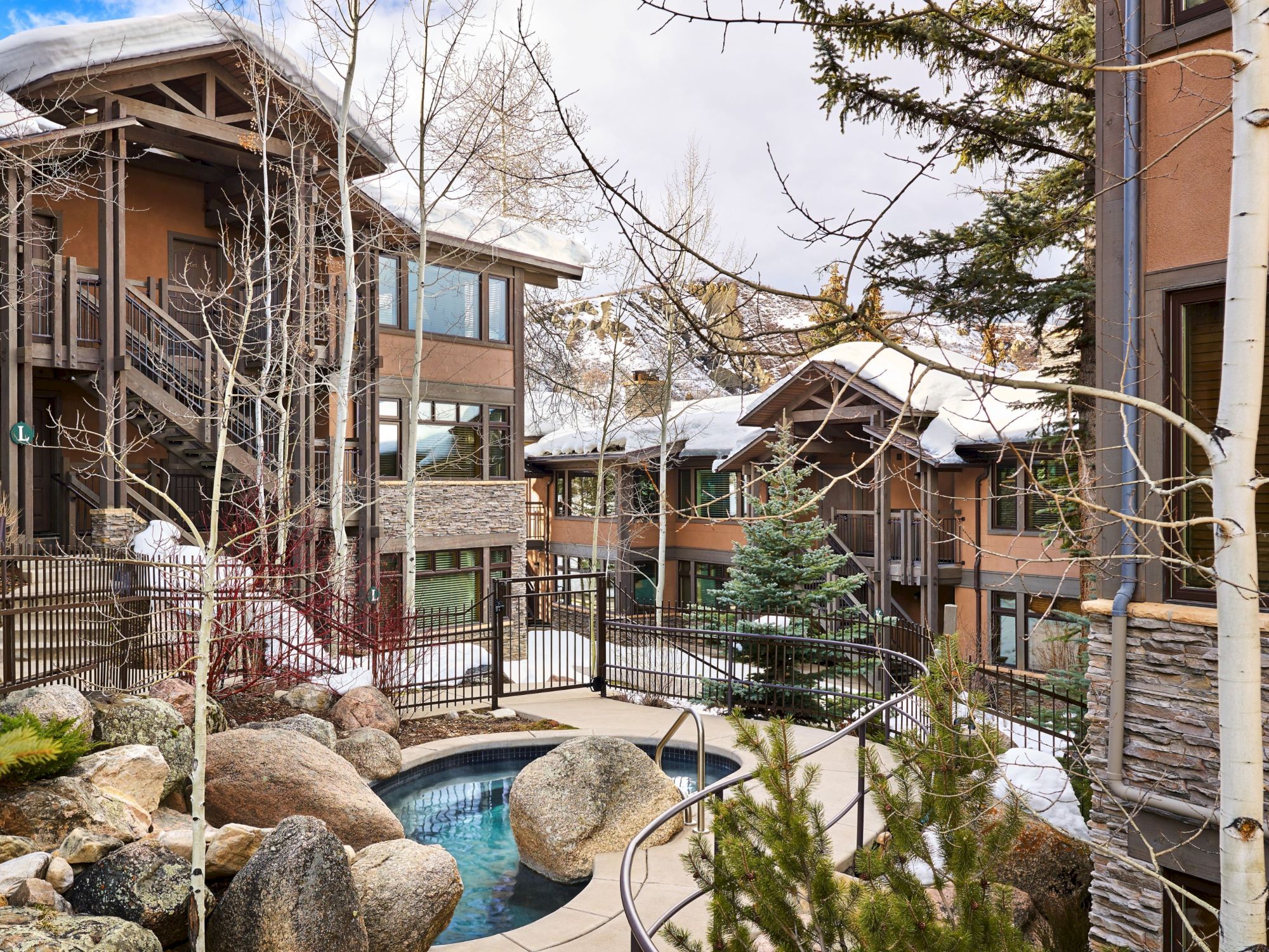 The image shows a winter resort courtyard with snow-covered buildings, a hot tub, and surrounding trees and rocks. It's serene and inviting.