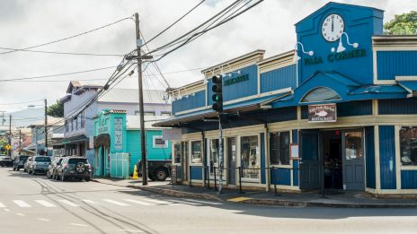A street view featuring colorful buildings, a clock on the blue building, parked cars, and power lines under a cloudy sky.