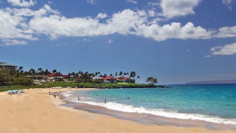 A sandy beach with turquoise waters, a few people swimming, buildings in the background, and palm trees under a partly cloudy sky.