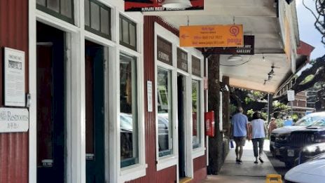 A street view showing a red building with a restaurant, outdoor signage, and a sidewalk with two people walking. Cars and trees are visible.