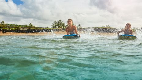 Two people are riding bodyboards in the ocean near a sandy beach with palm trees and cloudy skies.