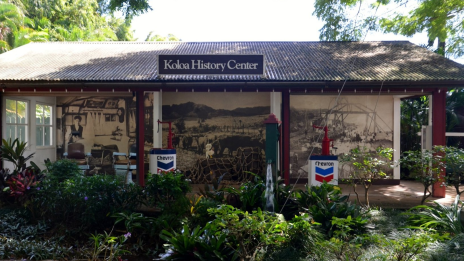 A building with a sign that reads "Koloa History Center," featuring Chevron gas pumps, historical photos, and lush greenery in the foreground.