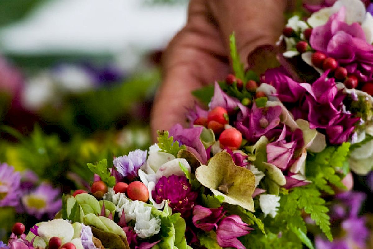 A person is holding a vibrant floral garland made of various colorful flowers and green foliage, including red berries, in their hand.