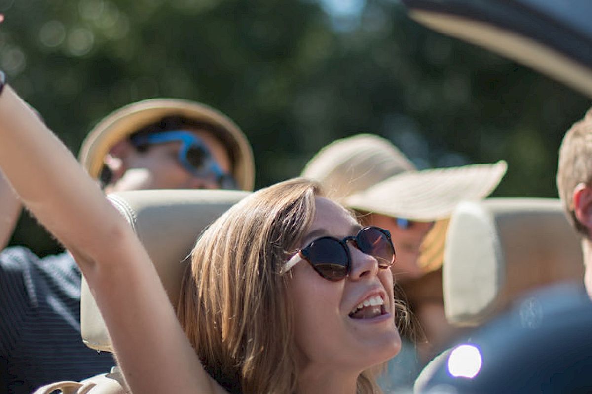 A group of young people wearing sunglasses is enjoying a ride in a convertible car, looking happy and carefree in the sunshine.