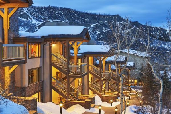 A snowy mountain scene features a well-lit lodge with multiple levels and bare trees, set against a backdrop of snow-covered hills at dusk.