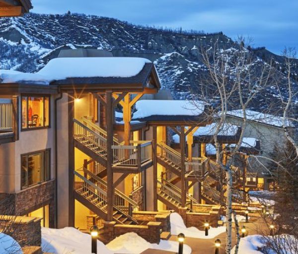A snowy mountain scene features a well-lit lodge with multiple levels and bare trees, set against a backdrop of snow-covered hills at dusk.