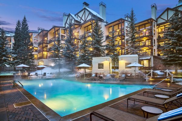 The image shows a cozy outdoor pool area surrounded by lounge chairs and umbrellas, with a snowy alpine lodge in the background.