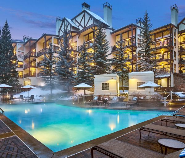 The image shows a cozy outdoor pool area surrounded by lounge chairs and umbrellas, with a snowy alpine lodge in the background.