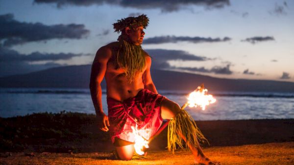 A person performs with fire on a beach at sunset, wearing traditional attire with a grass skirt and headpiece.