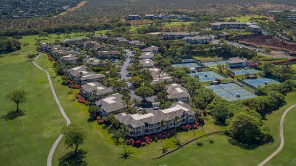 Aerial view of a resort-like complex with multiple white buildings, green lawns, palm trees, and tennis courts, set in a spacious, landscaped campus.