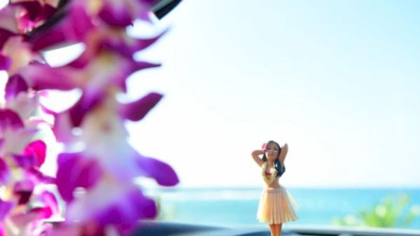 A hula girl figurine sits on a car dashboard with a blurred view of tropical flowers and the ocean in the background, under a clear blue sky.