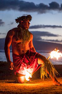 A person performs a fire dance on a beach at sunset, wearing traditional attire and surrounded by a scenic ocean backdrop.