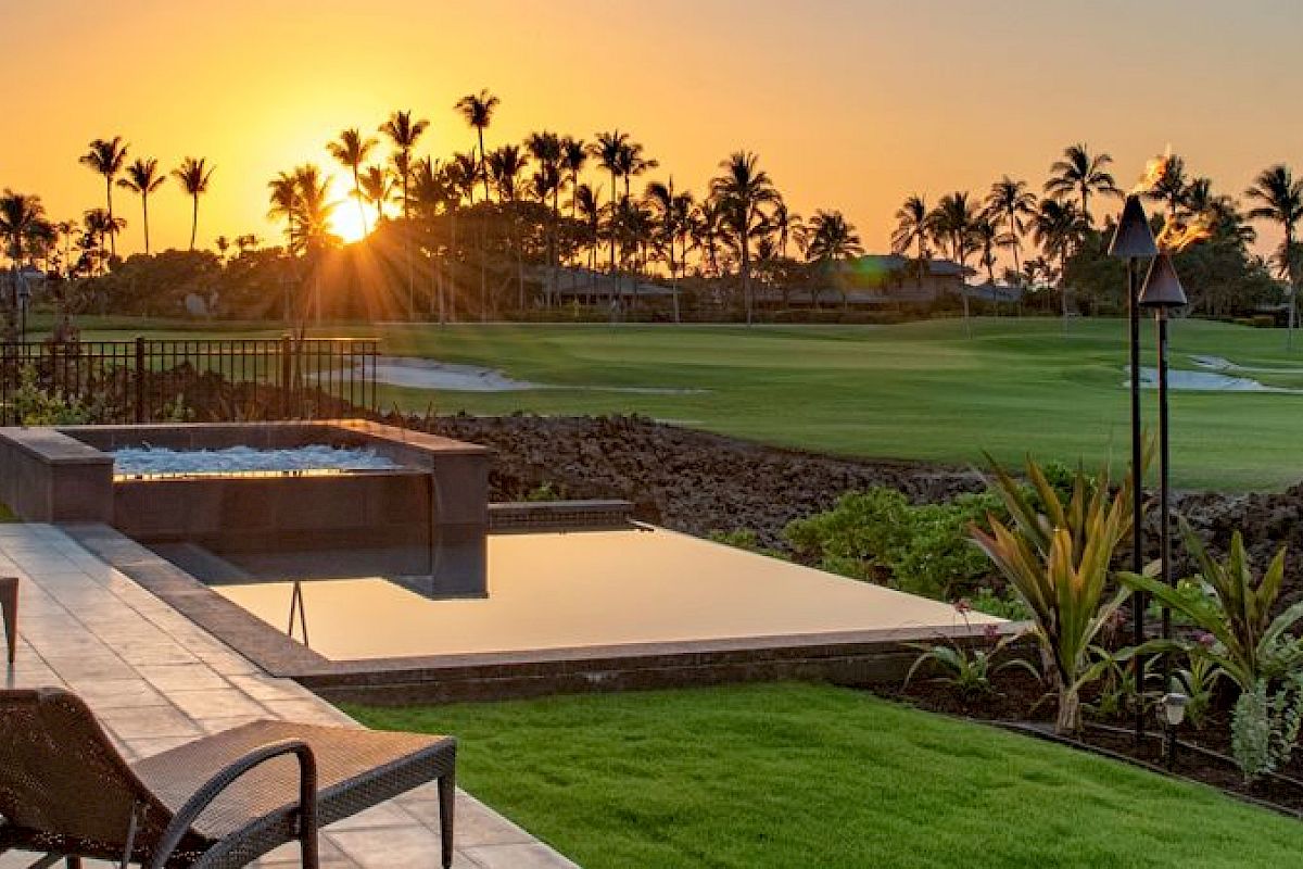 A beautiful sunset view from a patio featuring lounge chairs, an infinity pool, lush greenery, and a golf course surrounded by palm trees.