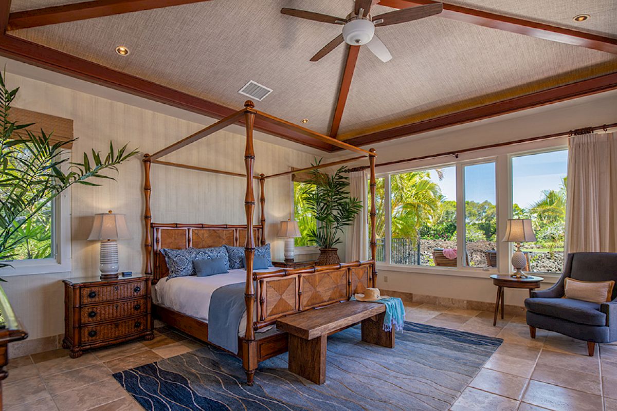 A spacious and well-lit bedroom with a four-poster bed, large windows, a ceiling fan, potted plants, and a chair, creating a cozy ambiance.