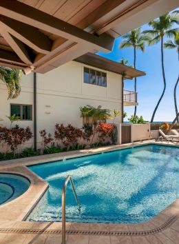 A sunny resort pool scene with a curved hot tub, palm trees, and lounge chairs along the deck, under a shaded building overhang, paradise.
