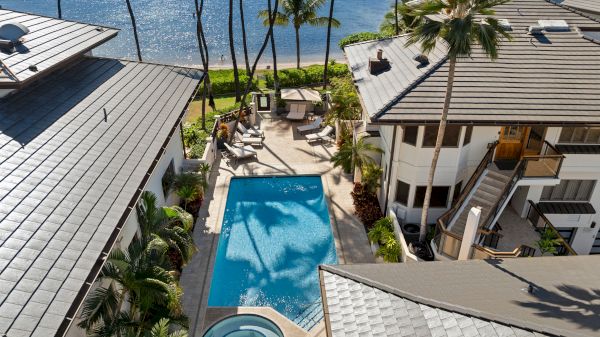 A sunny resort with a rectangular pool, circular spa, palm trees, and seaside views between two modern white buildings by the beach.