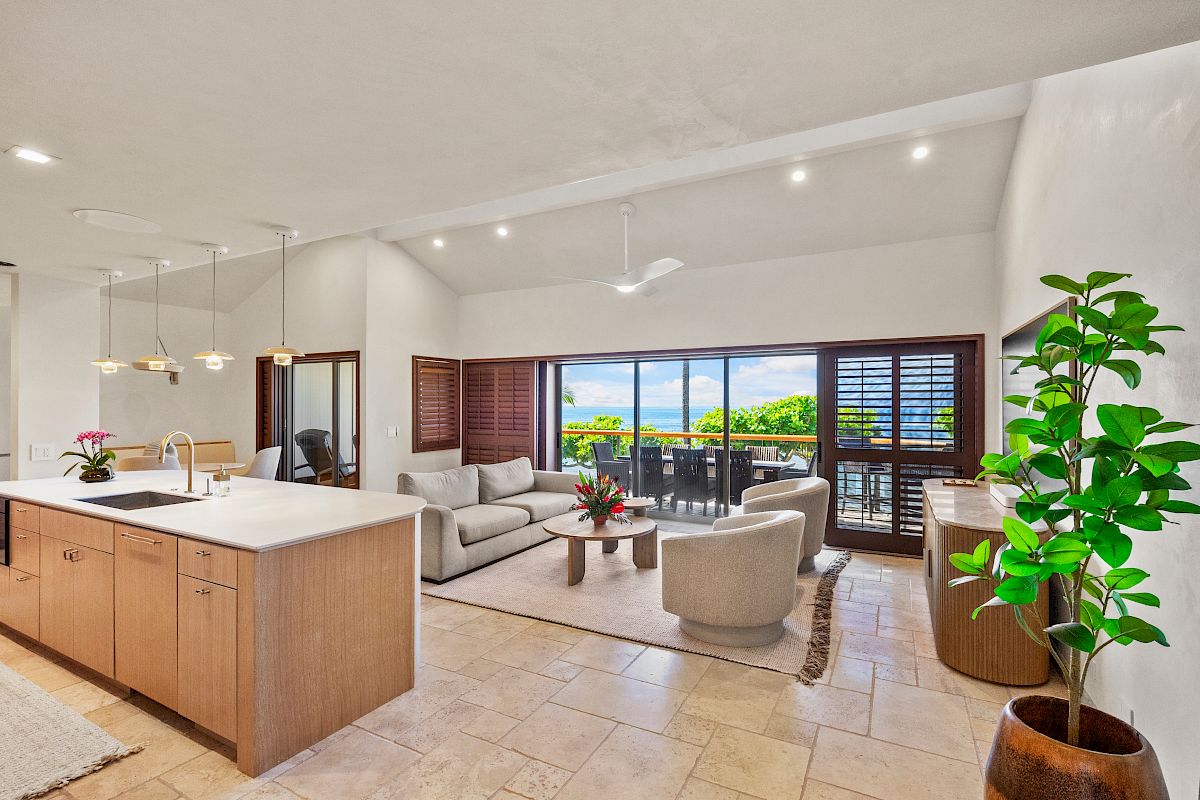 A bright, open-concept living area with a kitchen island, comfy seating, large glass doors to a balcony, and potted plants.