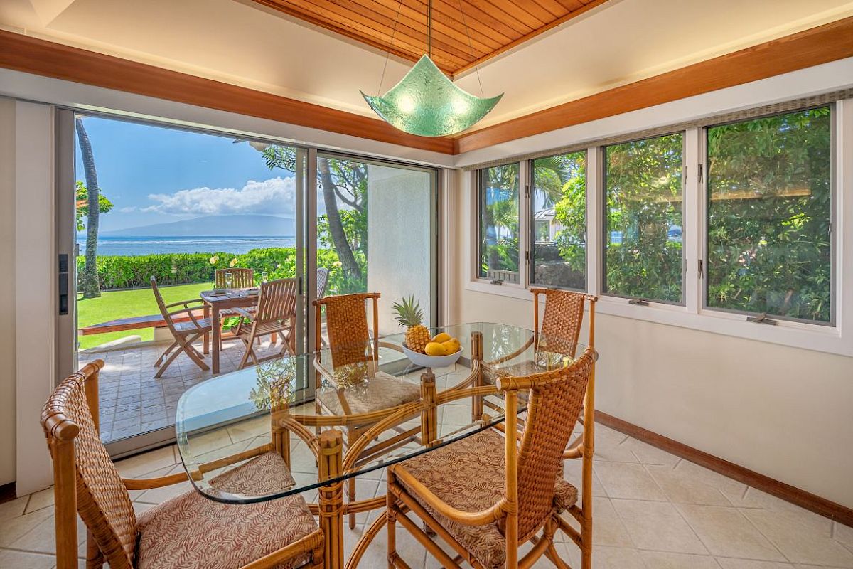 A bright dining room with a glass table, wicker chairs, large windows, and a view of an outdoor patio and lush greenery facing the ocean.