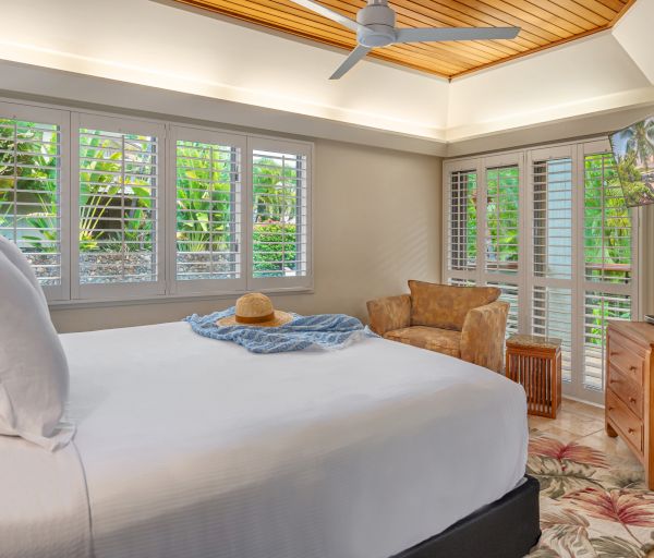 A cozy bedroom with a large bed, a ceiling fan, a TV, and large windows allowing abundant natural light, showing a tropical garden view outside.