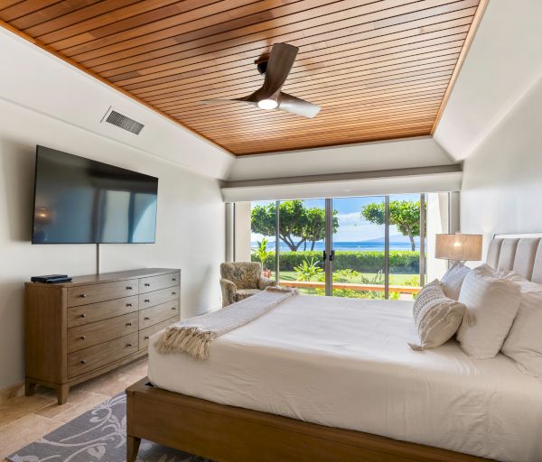 A bright hotel room with a wooden ceiling, two queen beds, a dresser with a wall TV, and a large window overlooking a sunny lawn and water.