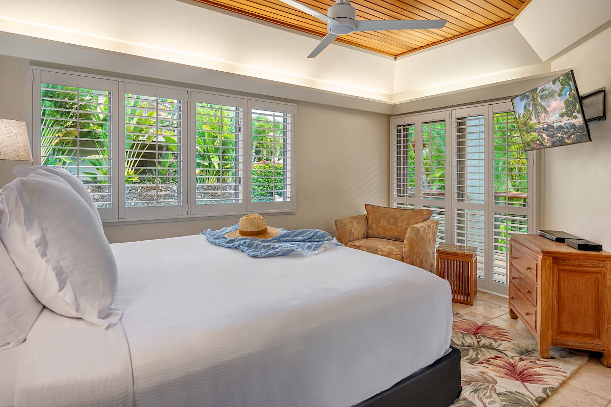 A cozy bedroom with a bed, hat, and scarf, adjacent to a window with blinds. There is a chair, TV, and tropical-themed décor. A door leads outside.