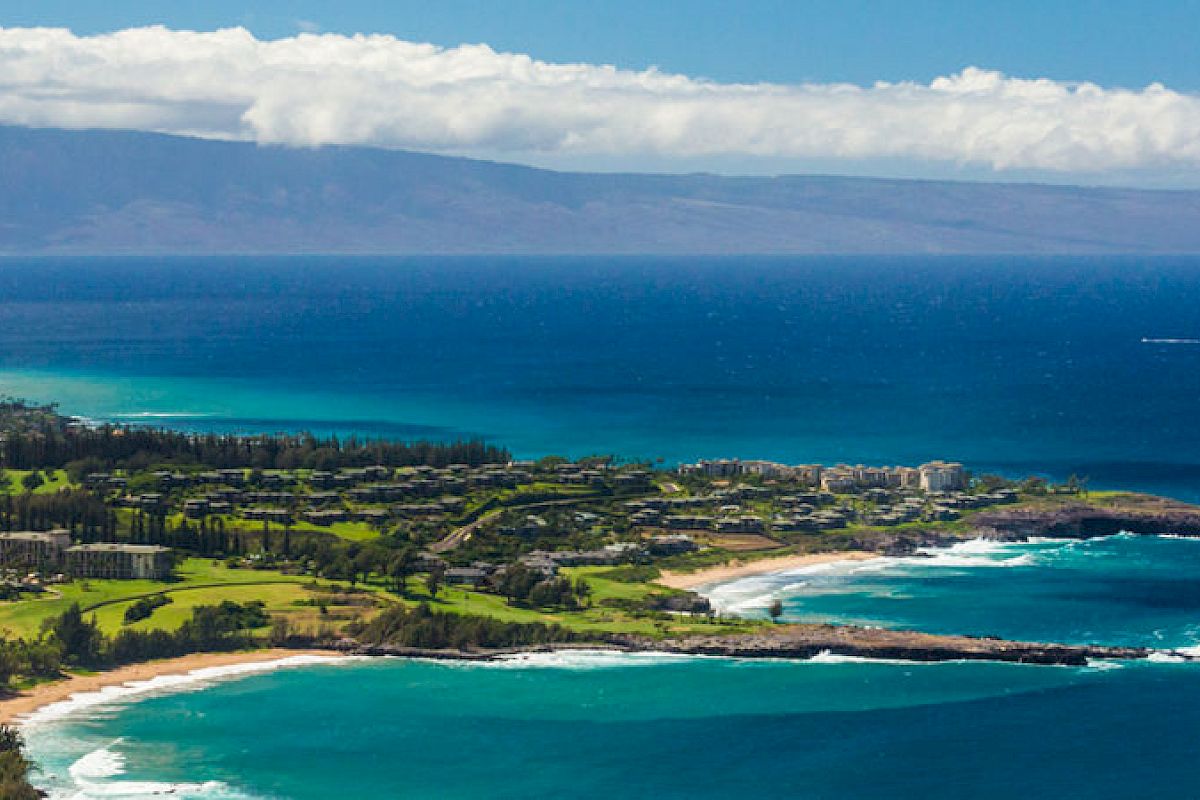 A coastal landscape featuring lush greenery, white sandy beaches, and turquoise ocean waters with a mountain range in the background under a bright sky.