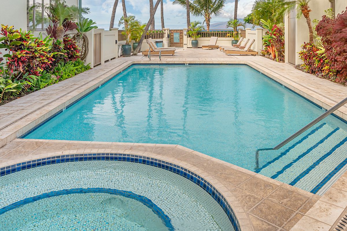 This image depicts a pristine swimming pool with adjacent hot tub surrounded by lush greenery and tropical plants on a tiled deck.