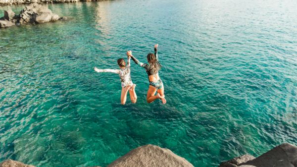 Two people jumping into clear turquoise water from rocks by the coast, mid-air with arms outstretched, sunny day.