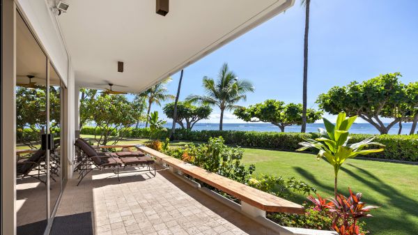 A sunny poolside patio with lounge chairs, glass doors, a manicured lawn, palm trees, and a view of the ocean in the distance, serene.