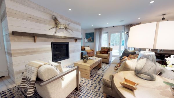 A cozy, modern living room with a fireplace, various seating, a coffee table, and a lamp. Large windows bring in natural light, enhancing the decor.