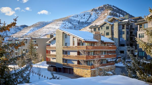 Snow-covered buildings with balconies, surrounded by evergreen trees, set against a mountainous backdrop under a blue sky with scattered clouds.
