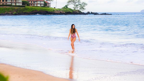 A person walking along a sandy beach with the ocean in the background and some buildings and trees in the distance.