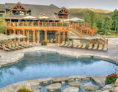 The image shows a luxurious outdoor pool area surrounded by lounge chairs and umbrellas, next to a large wooden lodge-style building with scenic greenery.