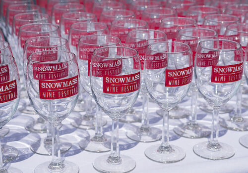 The image shows a table filled with wine glasses, each featuring a label for the Snowmass Wine Festival.