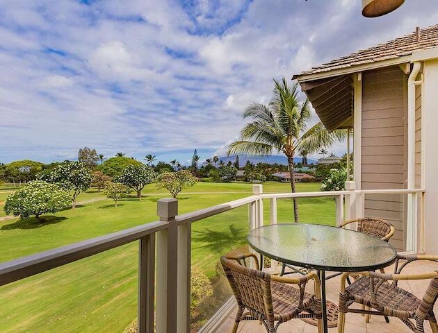 A balcony with a glass table and wicker chairs overlooks a lush golf course with palm trees under a partly cloudy sky.