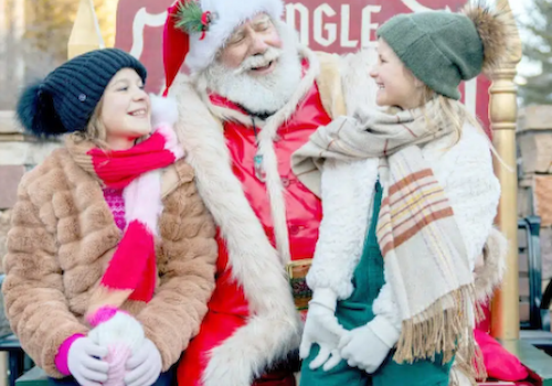 The image shows Santa Claus sitting with two smiling girls, enjoying a festive winter moment outdoors. Joy fills the scene.