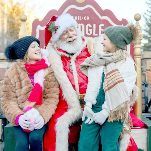 The image shows Santa Claus sitting with two smiling girls, enjoying a festive winter moment outdoors. Joy fills the scene.