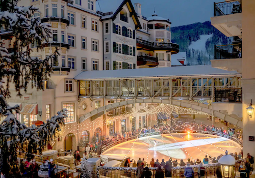 The image shows a lively ice skating rink in a winter setting, surrounded by charming buildings and a festive atmosphere with spectators.