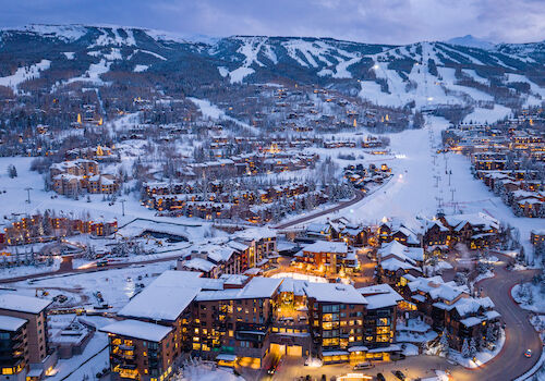The image shows a snowy mountain landscape with ski slopes, a cozy resort, and illuminated buildings at dusk. It's scenic and inviting.
