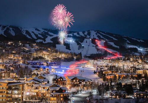 The image shows a night scene in a snowy village with colorful fireworks in the sky and illuminated buildings. It looks festive.