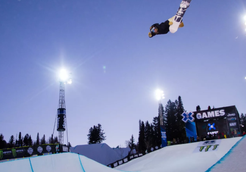 A snowboarder performs an aerial trick at a nighttime event, illuminated by bright lights, with spectators in the background.