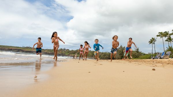 A group of kids runs joyfully along a sandy beach, splashing in the water under a cloudy sky filled with excitement and laughter.